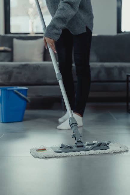 A person demonstrating surface cleaning in a modern living room, wearing a grey sweater, black pants, and white shoes, using a flat mop to clean a large, light-colored tile floor. The room features a grey fabric sofa with cushions, positioned against a large window allowing natural light to illuminate the space. A blue bucket with a mop wringer is placed nearby on the dark tiled floor. The scene emphasizes hygienic practices for residential cleaning, showcasing the process of deep cleaning and maintenance for tiled surfaces in a domestic setting, as promoted by Cleaners Edgware on their website page about Edgware house cleaning rates HA8 explained.
