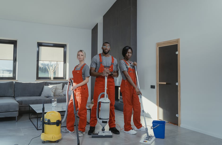 Three professional cleaners from Cleaners Edgware, dressed in grey shirts and red overalls, are positioned in a modern living room with light-colored walls and large windows. The female cleaner on the left holds a vacuum cleaner, while the male in the middle and the female on the right hold mops, all appearing ready for surface cleaning and deep cleaning tasks. The room features a grey fabric sofa with some cushions, a black glass coffee table, and a yellow vacuum in front of the cleaner on the left. The flooring is smooth, light-colored tile, and the lighting suggests natural daylight entering through the windows. The scene emphasizes thorough hygiene and maintenance work in a domestic setting, resonating with Edgware house cleaning rates HA8 explained, with Cleaners Edgware providing professional cleaning services to ensure surfaces are spotless and well-maintained.