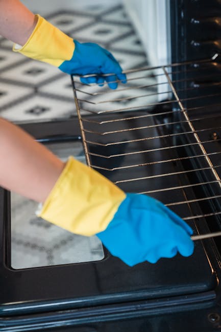 A woman engaged in surface cleaning on a white gas stove in a well-lit kitchen with natural light coming through a window. She wears a white t-shirt, a black apron, and yellow rubber gloves, holding a