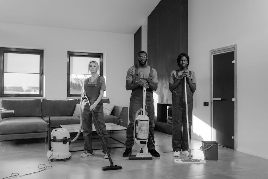 Three professional cleaners from Cleaners Edgware standing in a modern living room with a sofa, coffee table, and two large windows allowing natural light. They are equipped with cleaning tools: the f