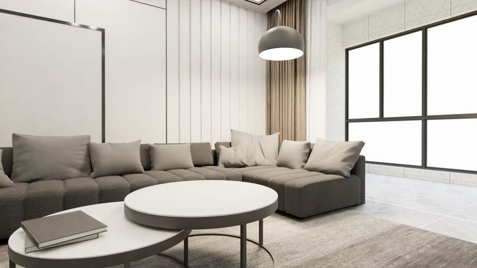 A spacious, well-lit living room with a series of large bay windows fitted with white blinds, allowing natural light to illuminate the beige carpeted floor which appears clean and tidy. The walls are 